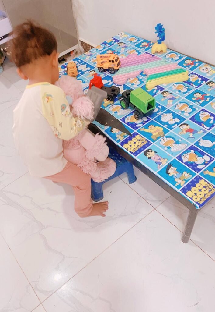 Toddler practicing empathy by placing a pink stuffed poodle on a blue stool to include it in play with toy trucks.