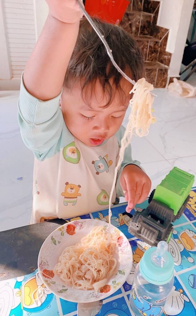 Toddler boy lifting spaghetti noodles high above his head with a fork while learning to eat.