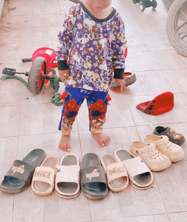 Toddler boy standing proudly next to a straight line of sandals and slides he arranged on the patio floor.