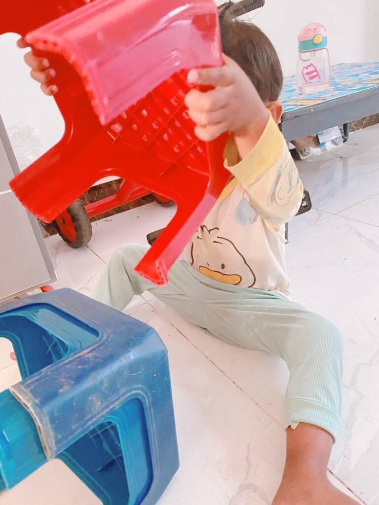 Toddler holding a red plastic stool up to his eyes and looking through the ventilation holes like a telescope.