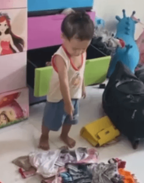 A toddler standing in a bedroom pointing proudly at a messy pile of clothes he pulled out of his dresser.