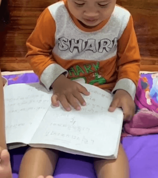 A toddler wearing an orange shark shirt sitting on a bed and pretend reading from an old notebook.