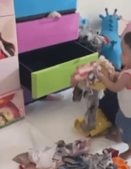 A toddler pulling folded clothes out of an open green plastic drawer to make a pile on the floor.