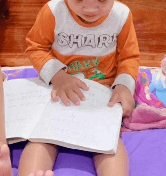 Toddler sitting calmly on a purple mattress reading a notebook while wearing his comfortable orange shirt.