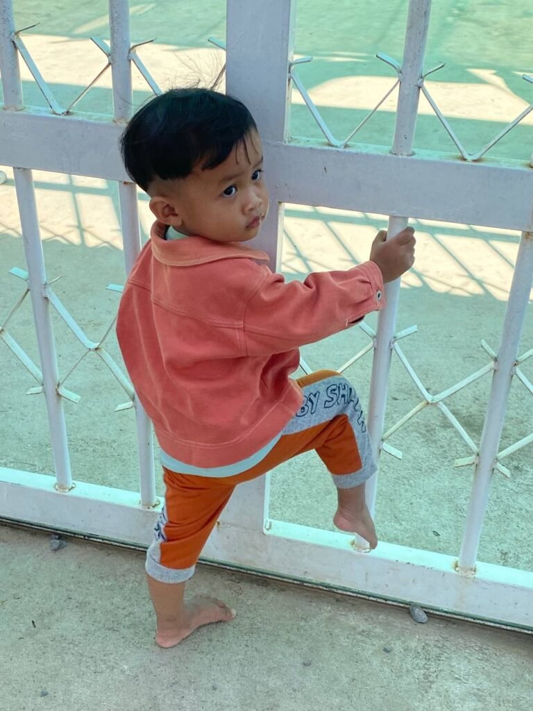 A toddler boy in an orange jacket climbing a white metal school gate barefoot, refusing to put sandals on.