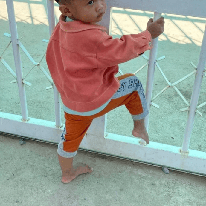 A toddler boy in an orange jacket climbing a white metal school gate barefoot, refusing to put sandals on.