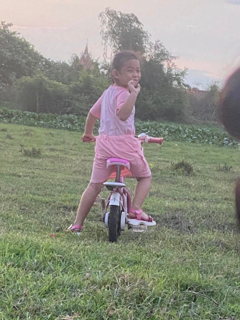 An 8-year-old girl smiling and looking back while riding a pink bicycle in a grassy field with a pagoda in the background.