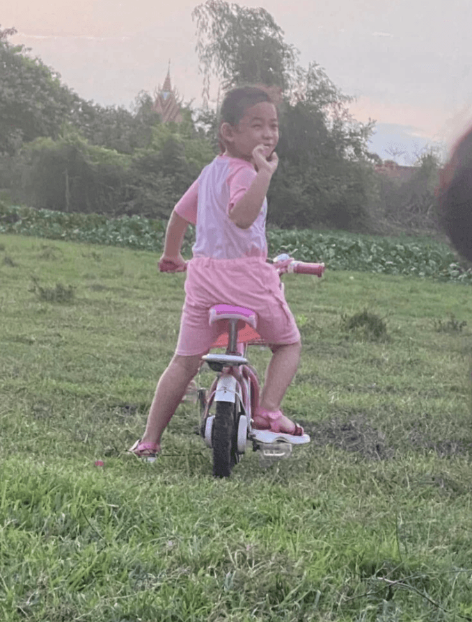 A young girl riding a pink bicycle in a grassy field with a Cambodian temple in the background after finishing her dinner.