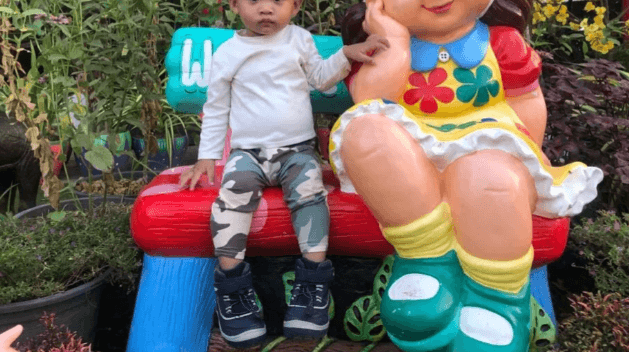 A toddler boy in a white shirt and camo pants sits on a colorful bench next to a large garden statue of a girl wearing a bright yellow flower dress, looking unimpressed.