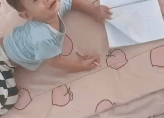 A smiling two-year-old boy lying on a peach blanket, looking up while holding a pencil and an open notebook.