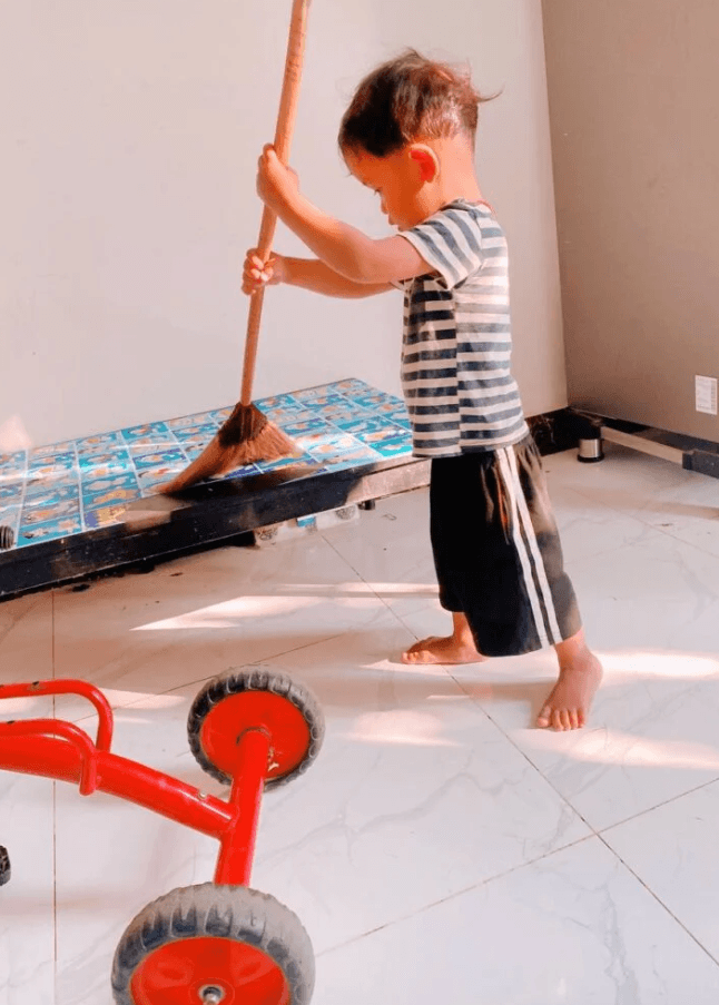 A serious toddler holding a broom with both hands, sweeping the floor next to a red tricycle.