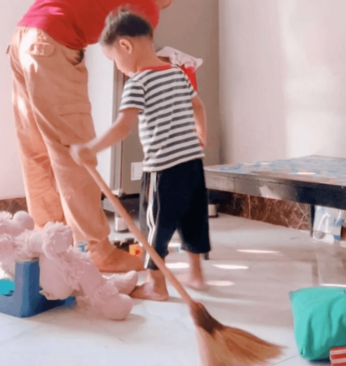 A toddler sweeping the floor next to a parent's feet, with toys scattered around the room.
