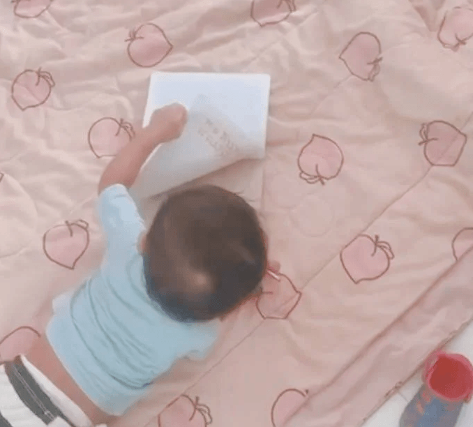A toddler grabbing the corner of a blank notebook page to turn it while lying on his stomach.
