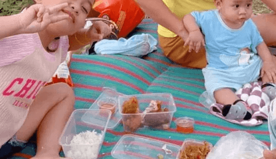 Two young children sitting on a striped picnic mat outside, happily eating rice and fried chicken directly from plastic storage containers.