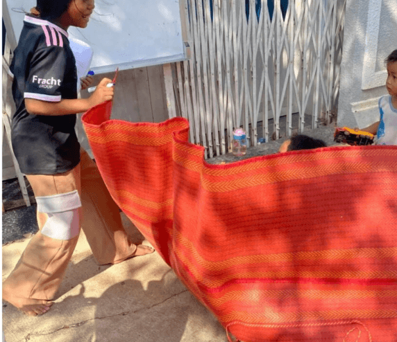 A gust of wind blowing a red woven mat up like a wall while a young girl laughs and a toddler holds a yellow dump truck.