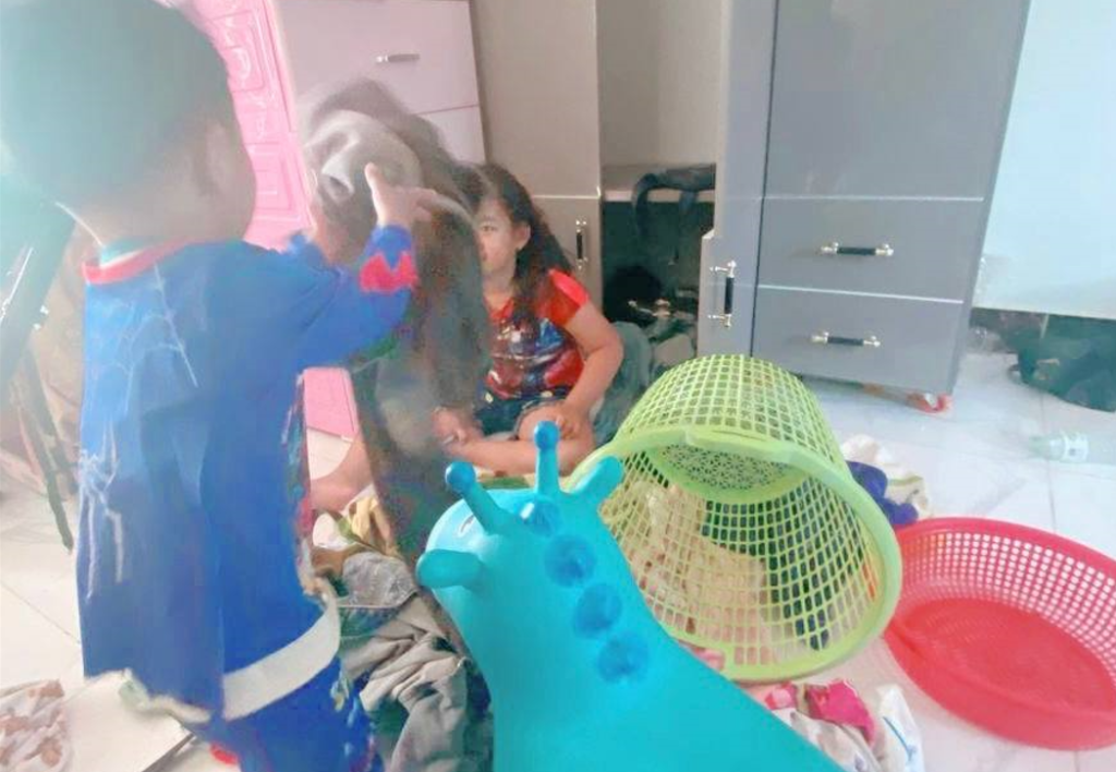 An 8-year-old girl sitting on the floor looking frustrated while her toddler brother in a Spider-Man suit plays.