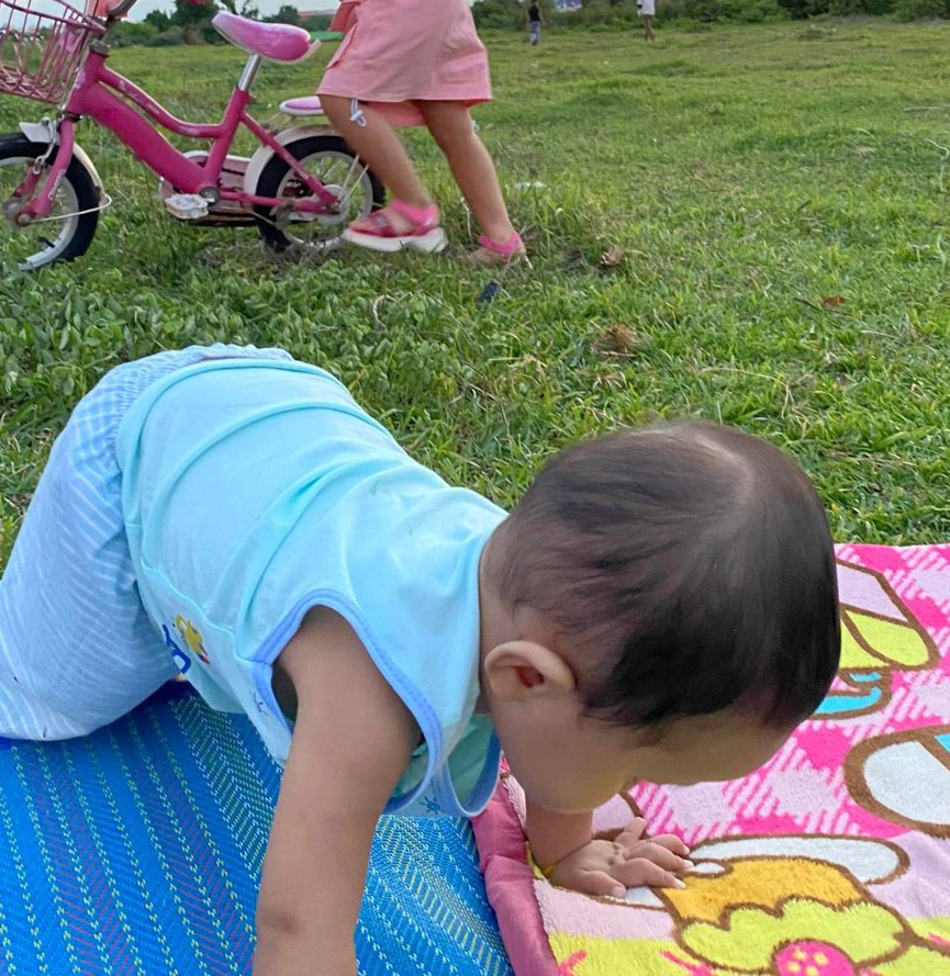A baby crawls on a colorful picnic mat while an older sibling rides a pink bike in a grassy field during a backyard safari.