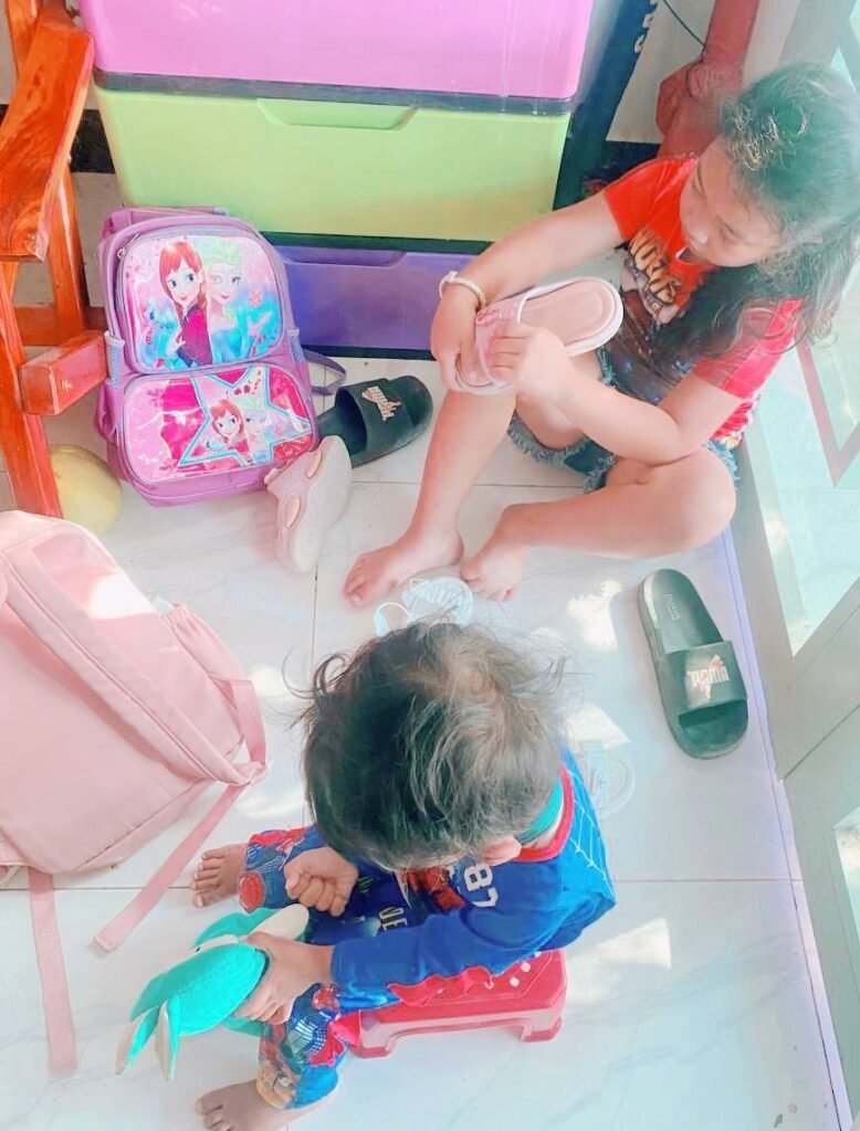 A young girl putting on pink sandals in slow motion next to a Frozen backpack by the door.