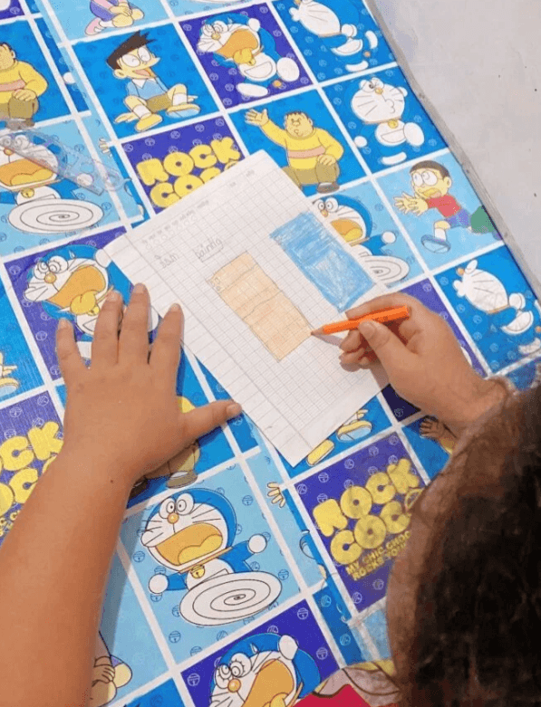 A child's hands coloring a chart with an orange colored pencil on graph paper on a Doraemon table.