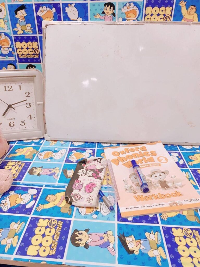 A simple homework setup featuring a white analog clock, whiteboard, and Oxford Phonics book on a blue table.