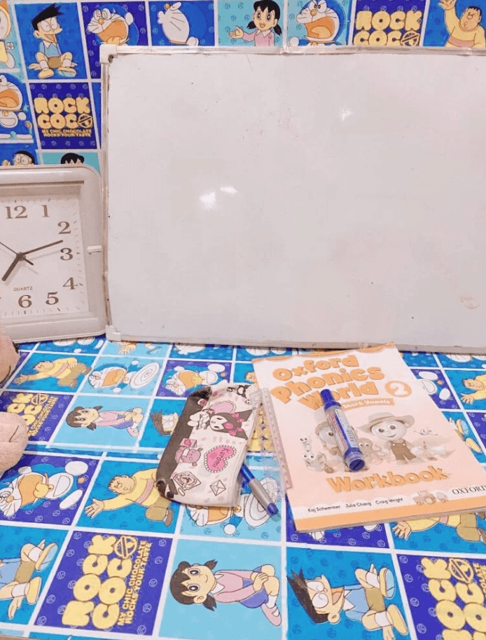 A low blue Doraemon table set up with a blank whiteboard, an analog clock, a workbook, and a marker.