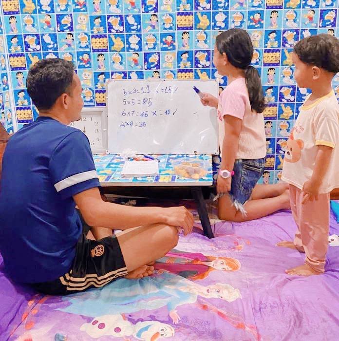 A father and his two children using a home classroom setup with a whiteboard against a wall decorated with blue cartoon decals.
