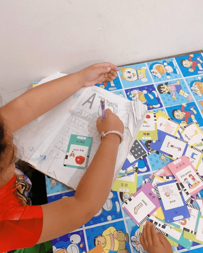 A child using a purple gel pen to trace the letter A in a workbook, with an apple flashcard placed on the page among scattered bilingual flashcards.