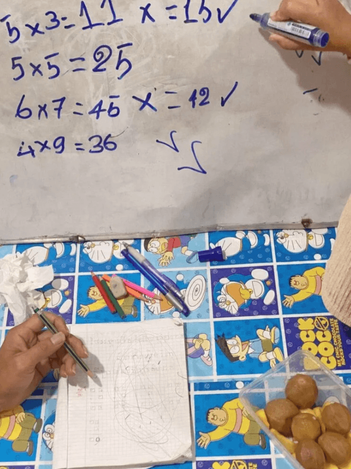A close-up of a whiteboard with corrected math mistakes, a notebook, and a container of round snacks on a blue Doraemon table.