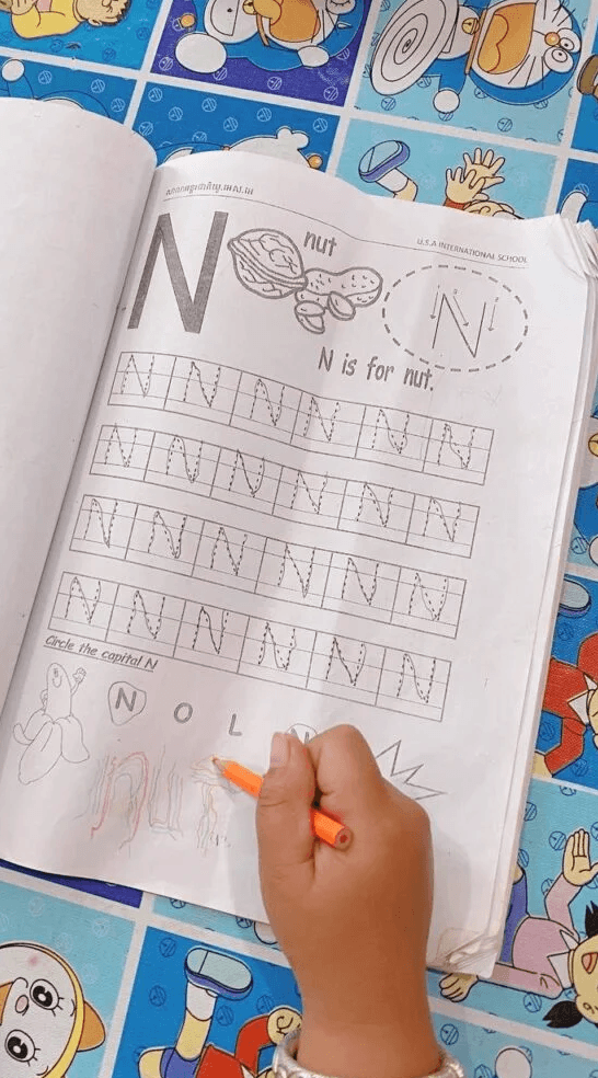 A child's hand using an orange colored pencil to trace the letter N on a workbook page with dotted lines.
