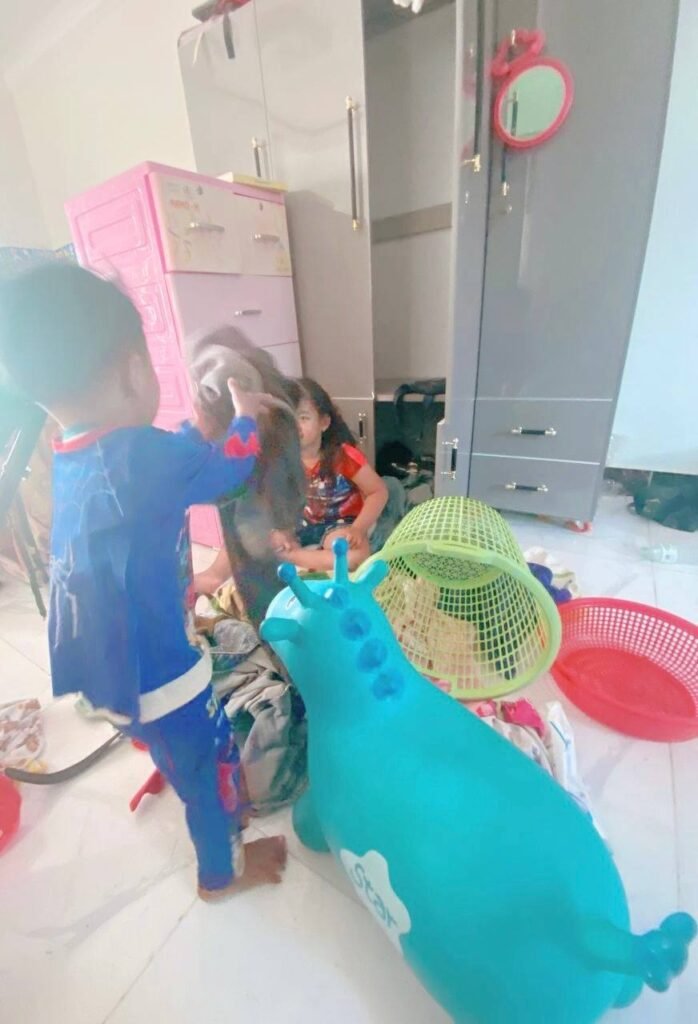An 8-year-old girl sitting on the floor looking frustrated while her toddler brother in a Spider-Man suit plays.