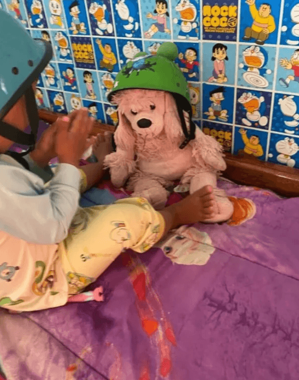 A toddler wearing a blue helmet sitting across from a pink stuffed poodle wearing a green helmet.