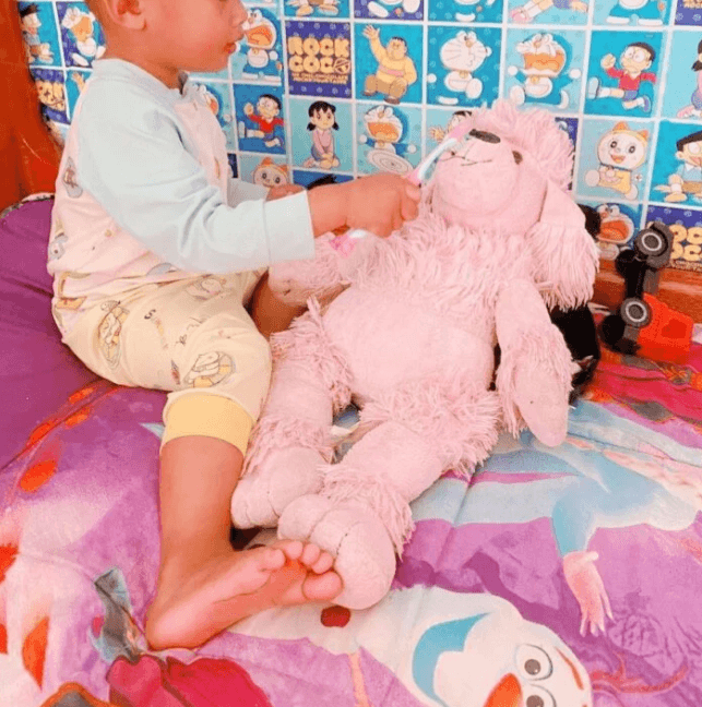 A toddler using a pink toothbrush to brush the teeth of a large pink stuffed poodle toy.