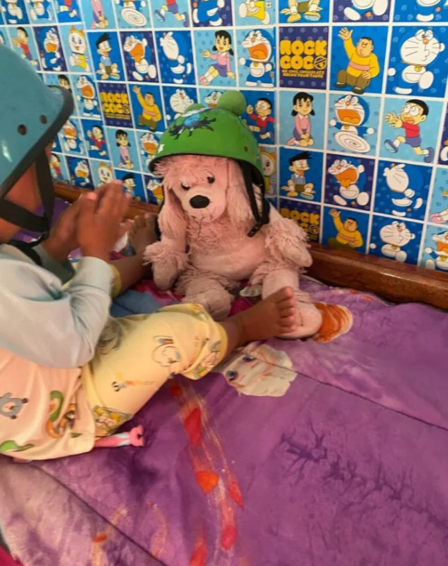 Pink stuffed poodle wearing a green bicycle helmet sitting on a bed.