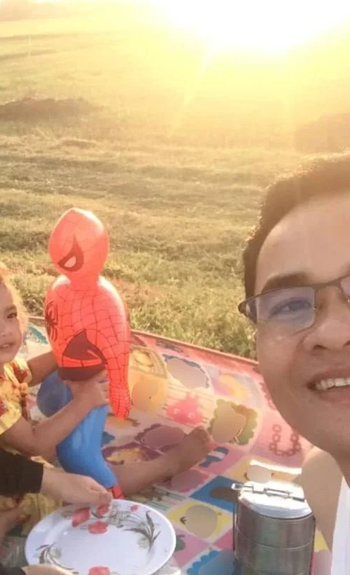 A father and daughter smile during a sunrise breakfast picnic in a grassy field, featuring a tiffin carrier and a giant inflatable Spiderman toy.