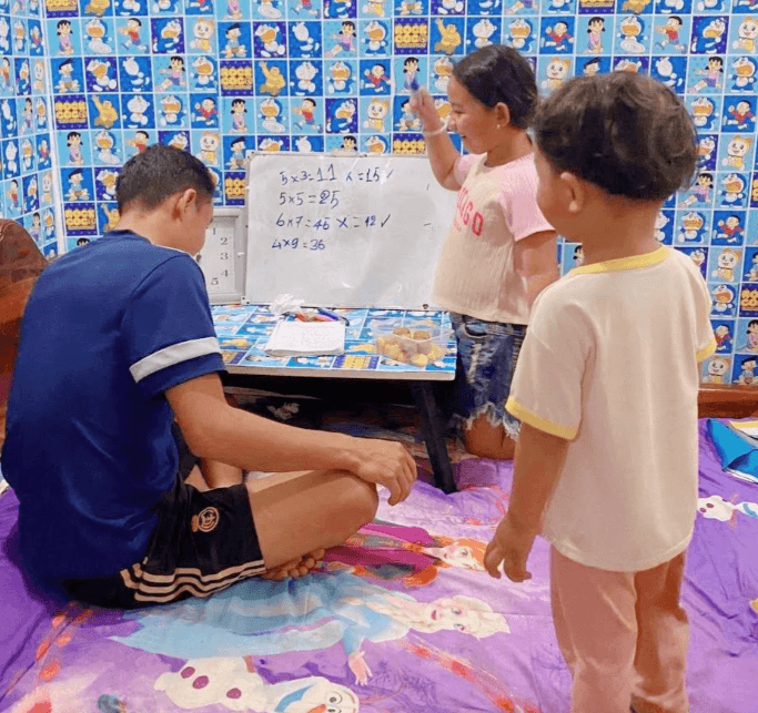 A father sitting on the floor while his 8-year-old daughter cheerfully corrects his math on a whiteboard, as her toddler brother watches.