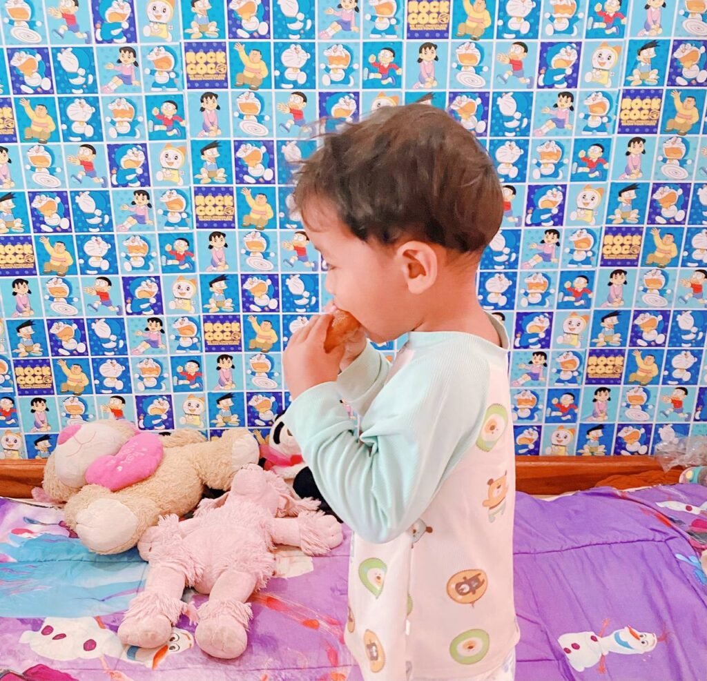 Side profile of a toddler boy eating a snack in a room with blue cartoon wallpaper.