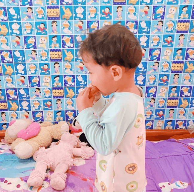 Side profile of a toddler boy eating a snack in a room with blue cartoon wallpaper.