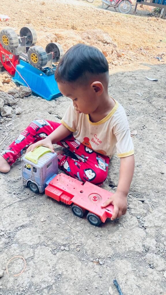 A toddler boy sitting in dry dirt, playing with a broken red and blue flatbed toy truck.