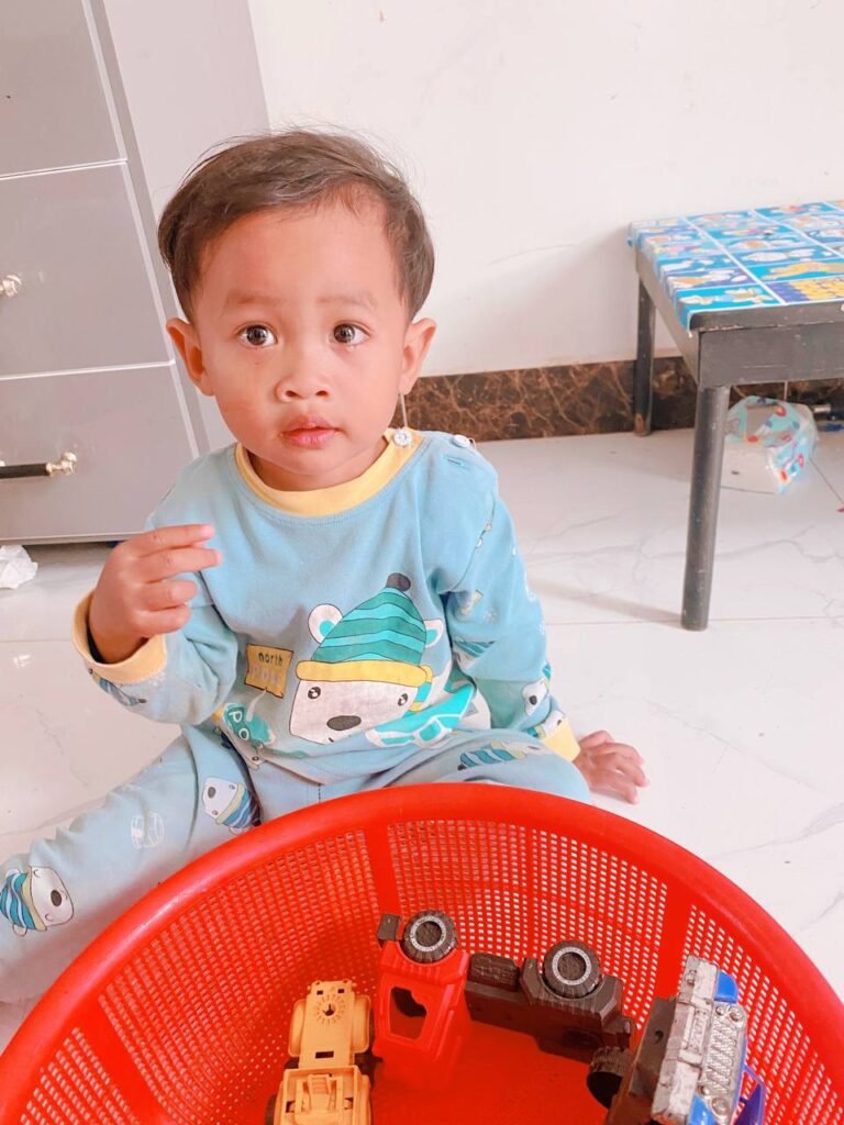 Cute toddler boy looking confused while sitting next to his red toy basket.