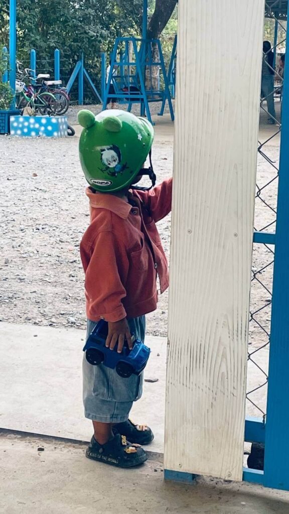A two-year-old boy in an orange jacket holding a blue toy truck while standing by a park gate.