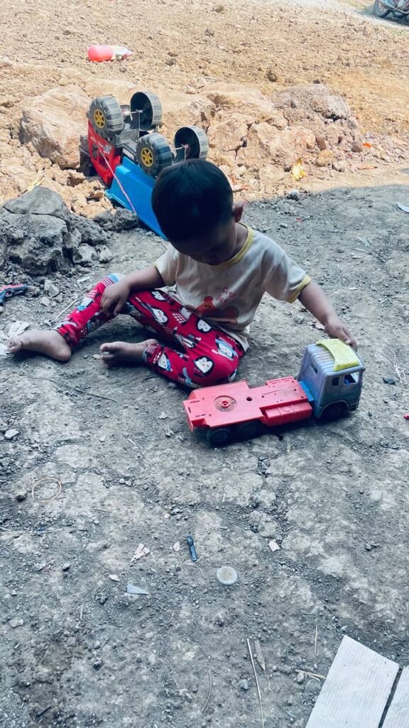 A toddler playing with a broken flatbed truck in the dirt, with a newer toy dump truck ignored in the background.