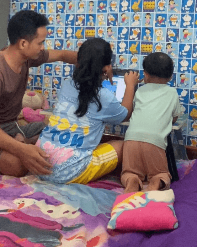 A toddler boy kneeling on a bed pointing at a laptop screen while his older sister types and their father watches.