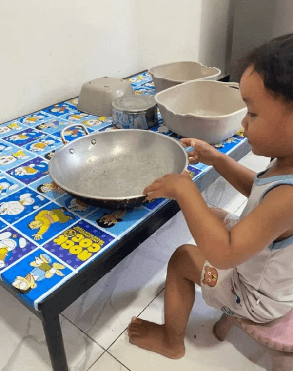 A toddler sitting at a small table playing with a large metal cooking pan and several plastic bowls.