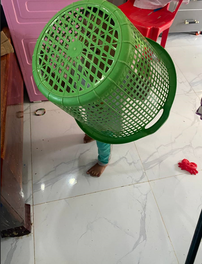 Toddler standing with a green plastic laundry basket covering their head and face.