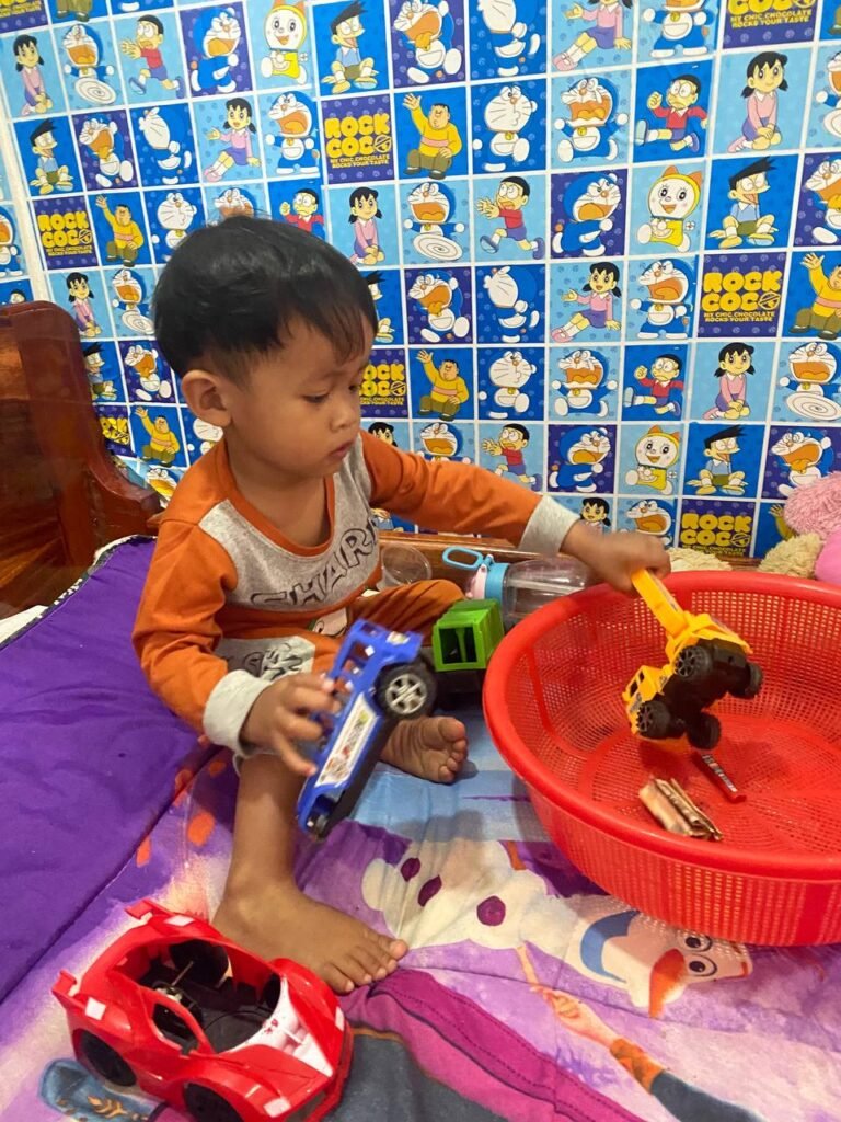 Toddler playing with toy trucks in a room covered with Doraemon wallpaper.