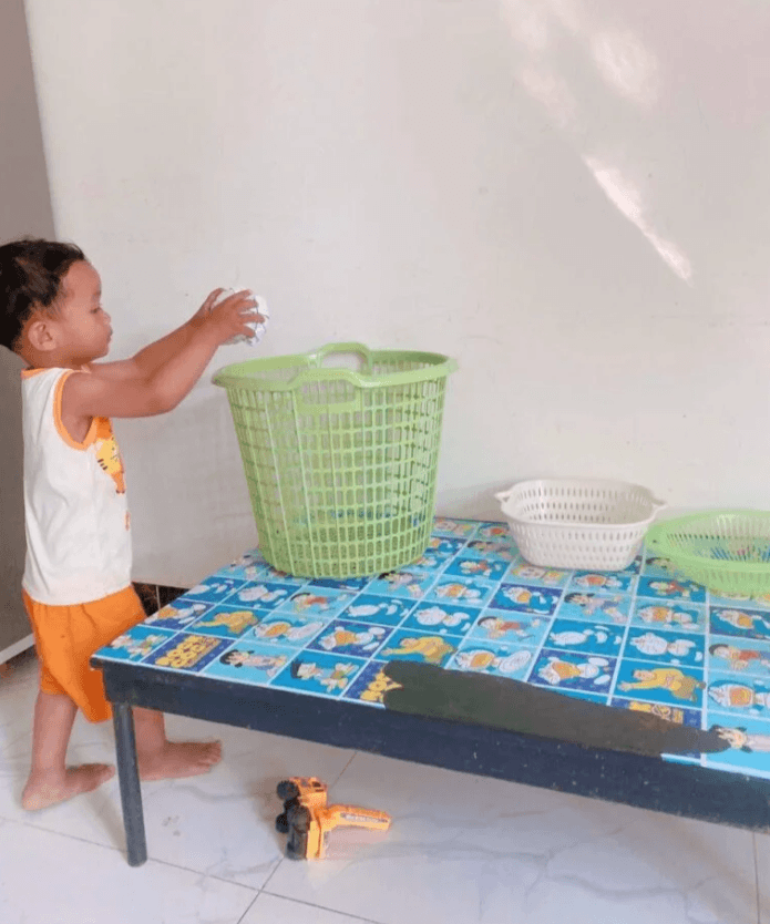 A child actively throwing a white paper ball into a large green plastic laundry basket, demonstrating household item play.