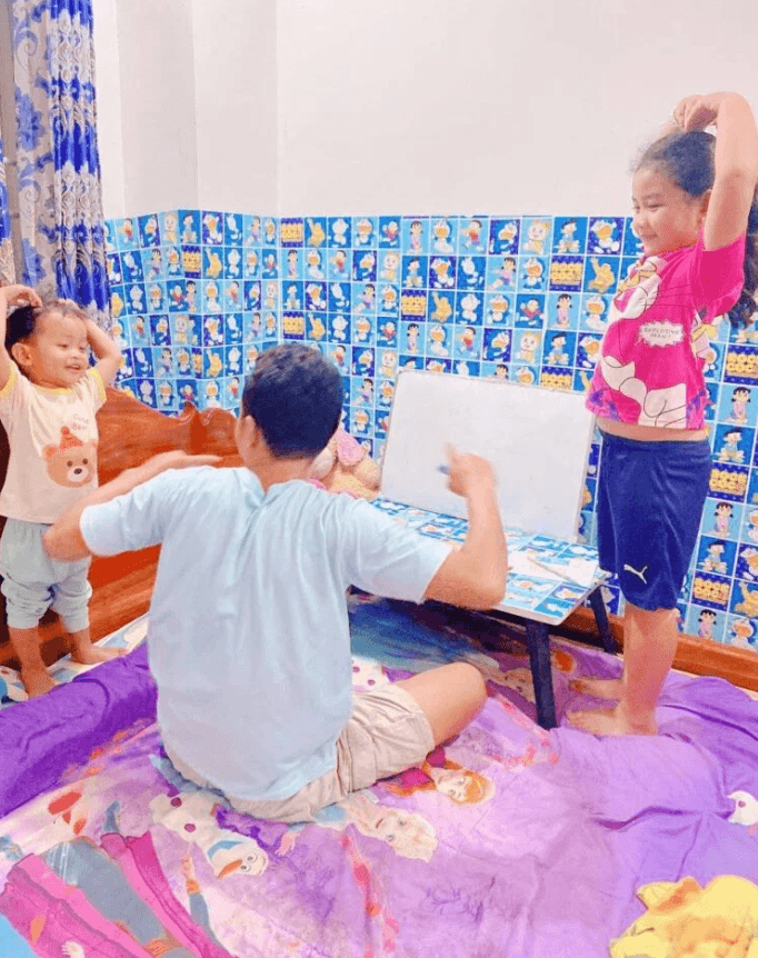 A father and toddler sitting on the floor while an 8-year-old girl stretches at a low Doraemon homework table.