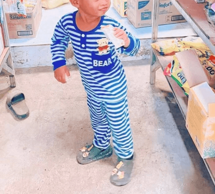 A toddler boy in striped pajamas standing in a store aisle holding a small snack box.