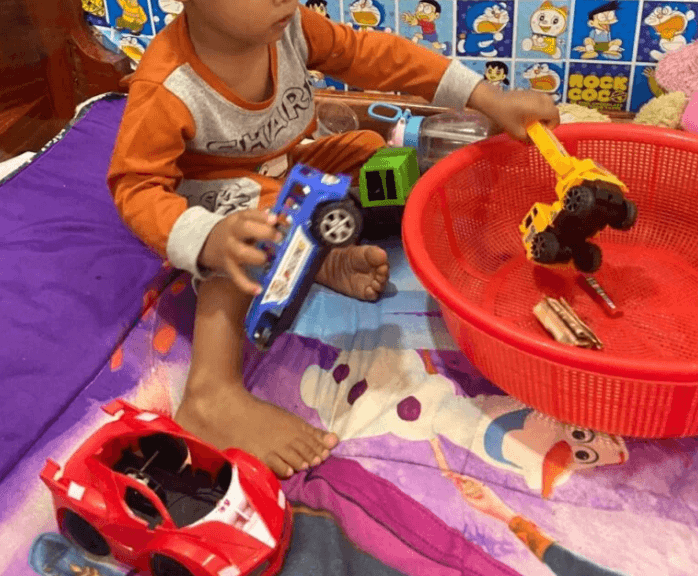 A toddler playing on a bed, putting a blue and a yellow toy truck into a red plastic basket.
