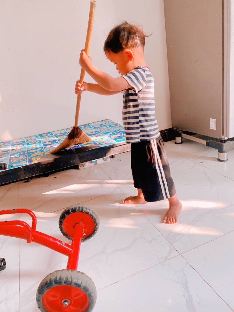 Toddler holding a real adult-sized broom and sweeping a play mat.